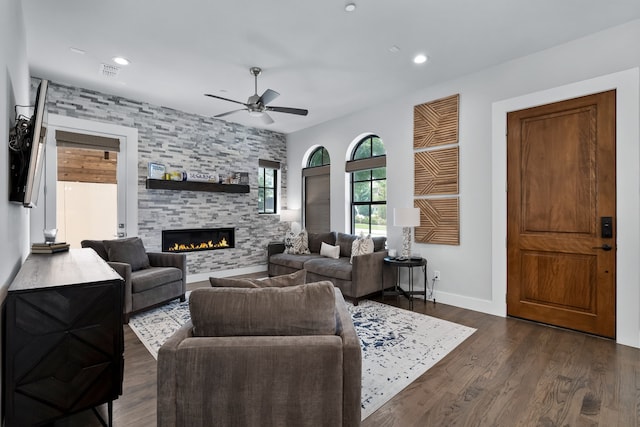 living room featuring dark wood-type flooring, ceiling fan, and a stone fireplace