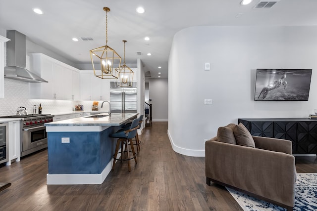 kitchen with pendant lighting, wall chimney range hood, a kitchen island with sink, white cabinetry, and high end appliances