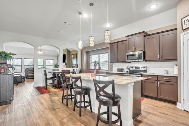 kitchen featuring decorative light fixtures, light stone counters, appliances with stainless steel finishes, light hardwood / wood-style flooring, and a kitchen island with sink