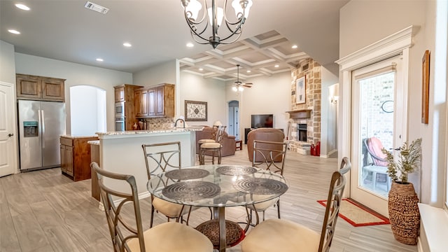 dining room with ceiling fan with notable chandelier, beamed ceiling, light hardwood / wood-style floors, coffered ceiling, and a fireplace