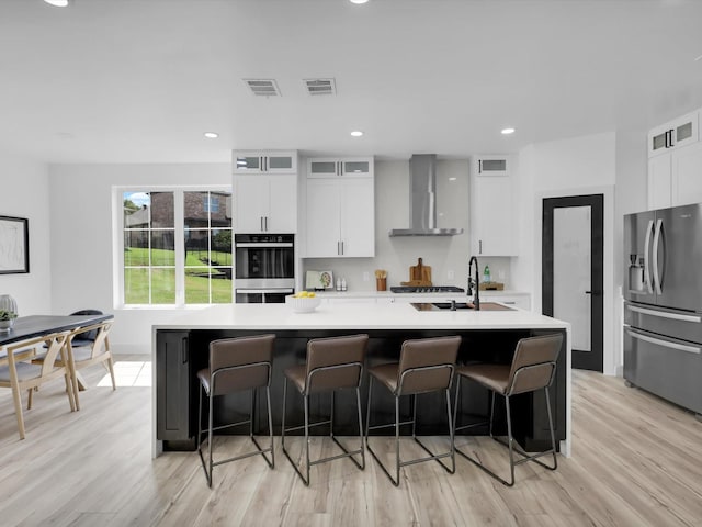 kitchen featuring light hardwood / wood-style flooring, stainless steel appliances, wall chimney exhaust hood, sink, and a center island with sink