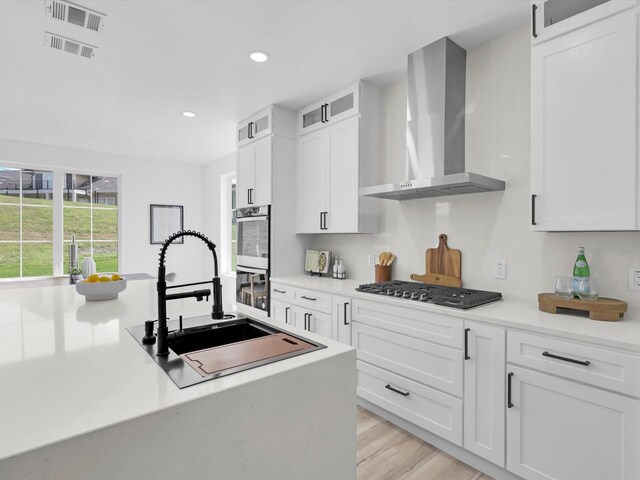 kitchen with white cabinetry, wall chimney range hood, sink, appliances with stainless steel finishes, and light hardwood / wood-style flooring