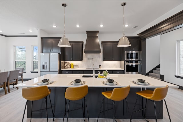 kitchen featuring appliances with stainless steel finishes, light hardwood / wood-style flooring, a center island with sink, hanging light fixtures, and custom range hood