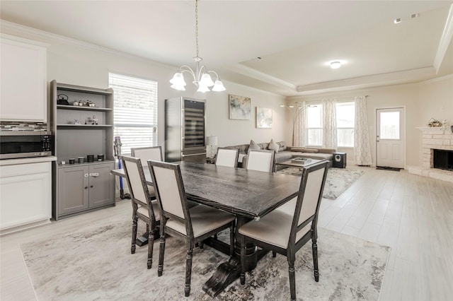 dining area with a chandelier, a stone fireplace, light wood finished floors, ornamental molding, and a tray ceiling