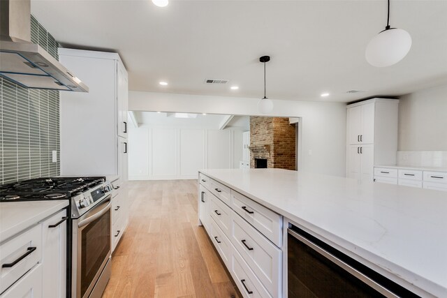 kitchen featuring wall chimney exhaust hood, decorative light fixtures, light hardwood / wood-style floors, white cabinetry, and stainless steel range with gas stovetop