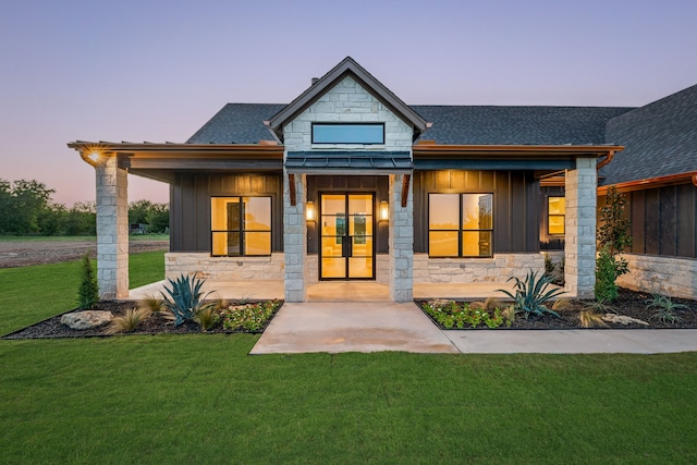 back of property at dusk with a yard, stone siding, board and batten siding, and roof with shingles