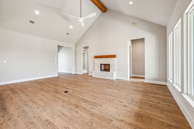 unfurnished living room with light wood-type flooring, ceiling fan, beamed ceiling, a stone fireplace, and high vaulted ceiling