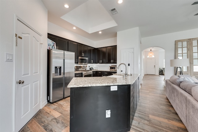 kitchen with appliances with stainless steel finishes, a center island with sink, sink, light stone counters, and hardwood / wood-style flooring