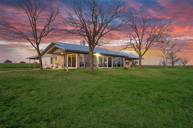 back of property at dusk featuring metal roof, a lawn, a chimney, and a sunroom