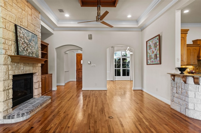 unfurnished living room with ceiling fan, a raised ceiling, a stone fireplace, light hardwood / wood-style flooring, and ornamental molding