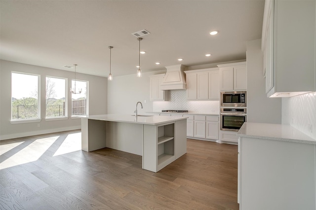 kitchen featuring light wood-type flooring, stainless steel appliances, custom exhaust hood, white cabinets, and a kitchen island with sink