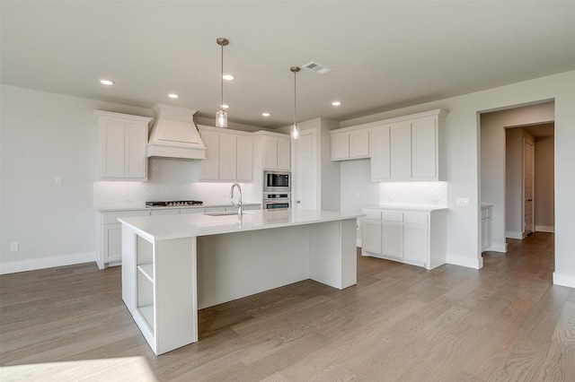 kitchen with an island with sink, appliances with stainless steel finishes, premium range hood, white cabinetry, and light wood-type flooring