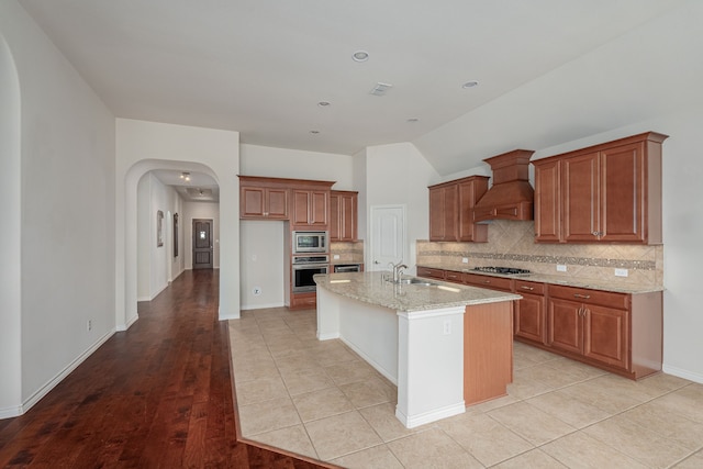 kitchen with custom exhaust hood, sink, a center island with sink, light hardwood / wood-style floors, and appliances with stainless steel finishes