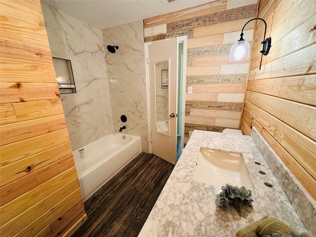 bathroom with wood-type flooring, wooden walls, vanity, and tiled shower / bath