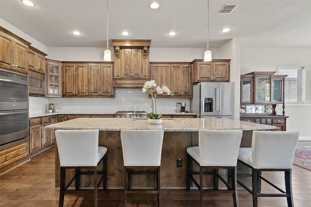 kitchen featuring a center island with sink, appliances with stainless steel finishes, dark hardwood / wood-style flooring, decorative light fixtures, and light stone countertops