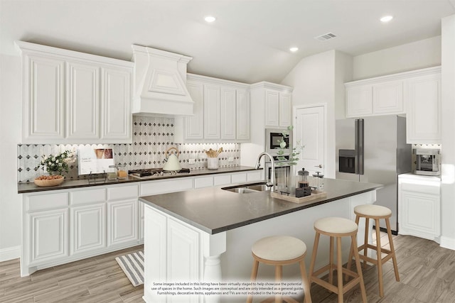 kitchen with sink, appliances with stainless steel finishes, white cabinetry, premium range hood, and an island with sink