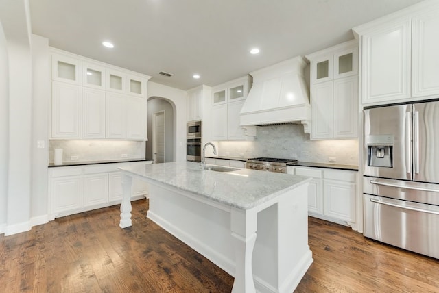 kitchen with sink, white cabinetry, appliances with stainless steel finishes, custom range hood, and a kitchen island with sink