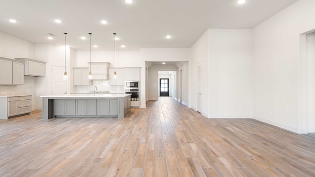 kitchen featuring an island with sink, hanging light fixtures, oven, built in microwave, and light wood-type flooring