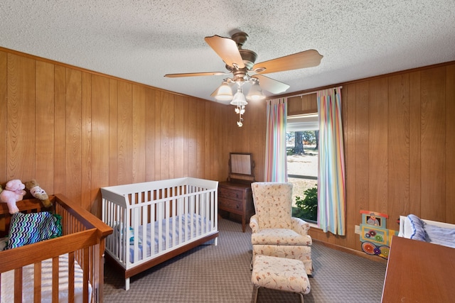 bedroom featuring ceiling fan, a crib, wooden walls, carpet, and a textured ceiling