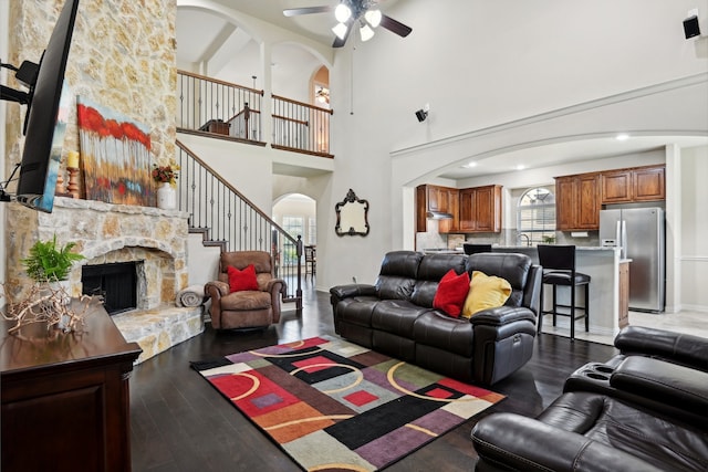 living room with ceiling fan, a towering ceiling, a stone fireplace, sink, and wood-type flooring