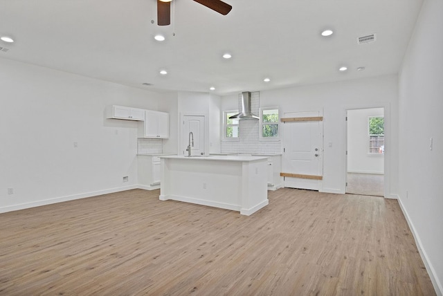 kitchen with white cabinetry, an island with sink, light hardwood / wood-style floors, wall chimney exhaust hood, and decorative backsplash