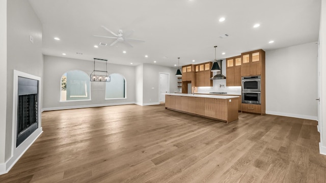 kitchen featuring wall chimney range hood, double oven, hardwood / wood-style floors, decorative light fixtures, and a large island with sink