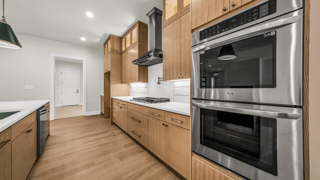 kitchen featuring wall chimney range hood, appliances with stainless steel finishes, hanging light fixtures, tasteful backsplash, and light hardwood / wood-style floors