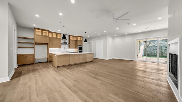 kitchen featuring wall chimney range hood, ceiling fan, hanging light fixtures, a large island with sink, and light wood-type flooring