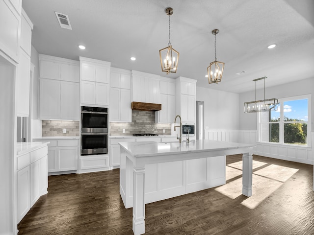 kitchen with double oven, a kitchen island with sink, white cabinets, and gas stovetop