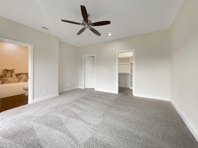 unfurnished bedroom featuring ceiling fan, a walk in closet, ensuite bathroom, and dark colored carpet