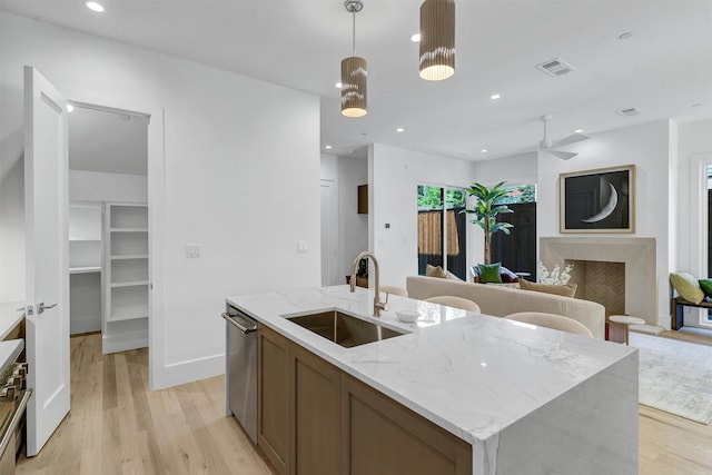 kitchen with stainless steel dishwasher, sink, light stone counters, an island with sink, and light wood-type flooring