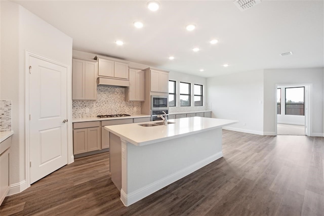 kitchen featuring appliances with stainless steel finishes, sink, decorative backsplash, a kitchen island with sink, and dark wood-type flooring