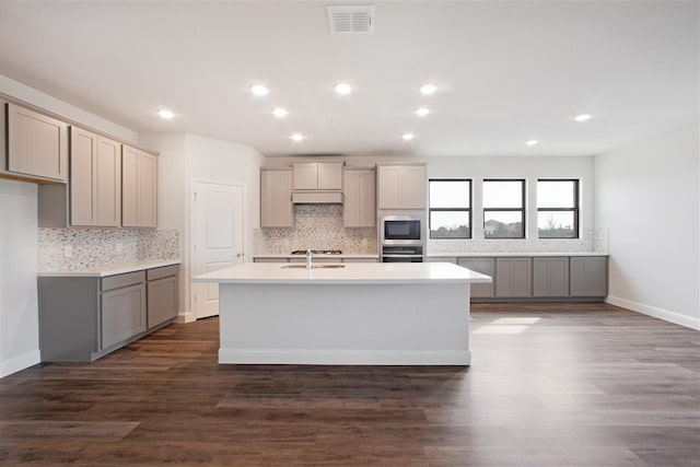 kitchen featuring stainless steel oven, wood-type flooring, an island with sink, black microwave, and gray cabinetry