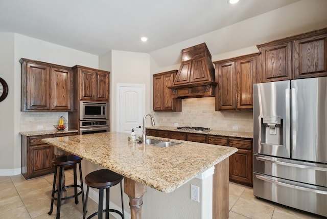 kitchen with custom range hood, tasteful backsplash, appliances with stainless steel finishes, sink, and a center island with sink