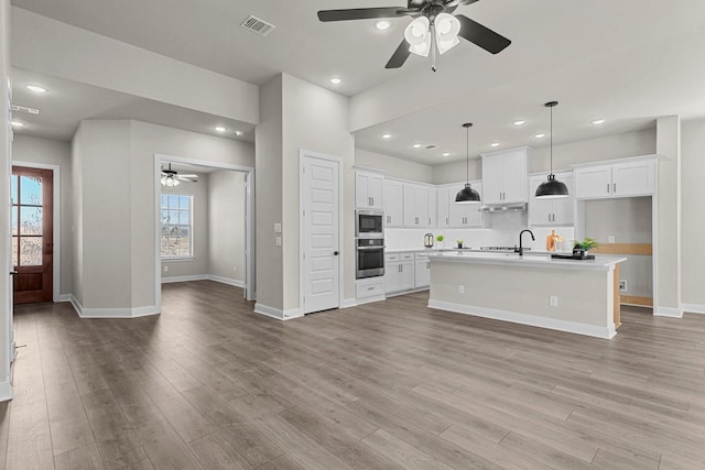 kitchen featuring white cabinets, hanging light fixtures, stainless steel appliances, a center island with sink, and light wood-type flooring