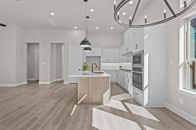 kitchen featuring appliances with stainless steel finishes, a kitchen island with sink, white cabinets, decorative light fixtures, and light wood-type flooring