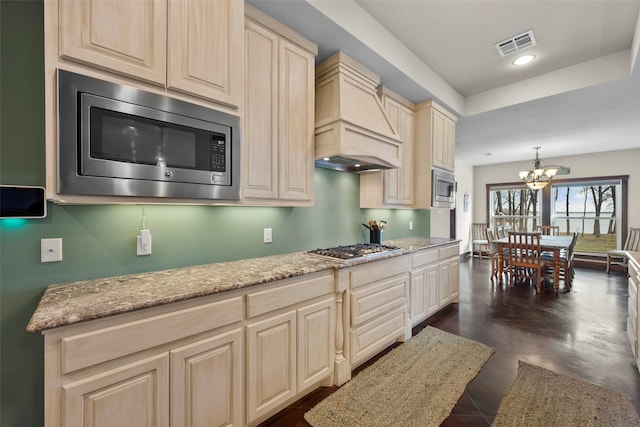 kitchen with an inviting chandelier, stainless steel appliances, hanging light fixtures, light stone counters, and premium range hood