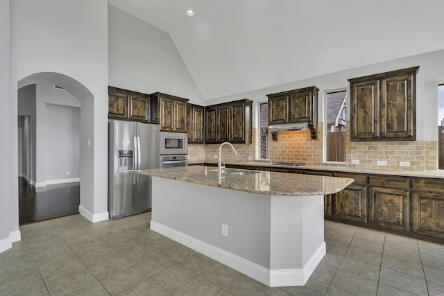 kitchen with stainless steel appliances, light stone countertops, an island with sink, and dark brown cabinets