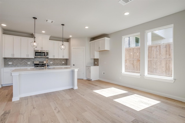 kitchen with appliances with stainless steel finishes, an island with sink, pendant lighting, and light hardwood / wood-style floors