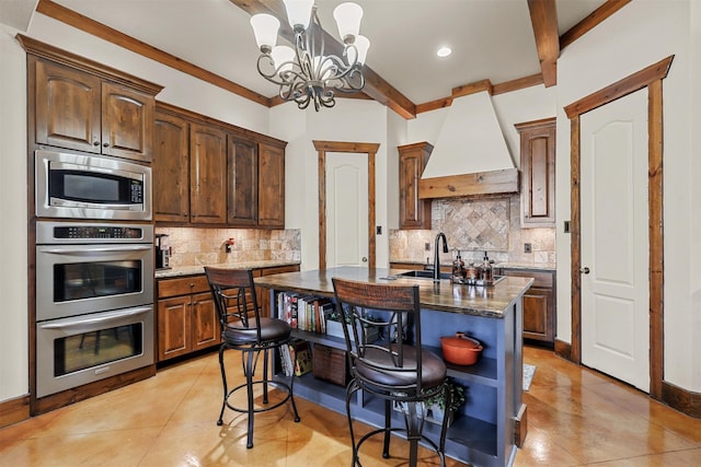 kitchen featuring appliances with stainless steel finishes, dark stone counters, an inviting chandelier, premium range hood, and a center island with sink