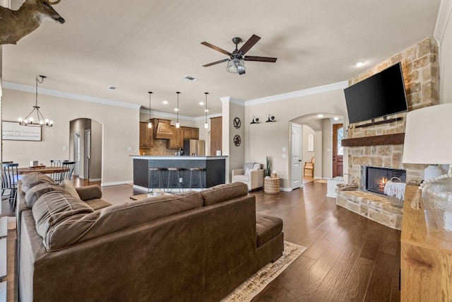 living room featuring ornamental molding, a stone fireplace, dark wood-type flooring, and ceiling fan with notable chandelier