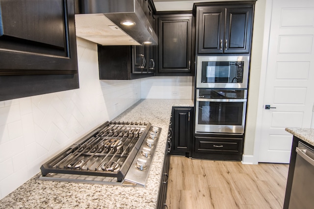 kitchen with wall chimney exhaust hood, light wood-type flooring, tasteful backsplash, light stone countertops, and appliances with stainless steel finishes