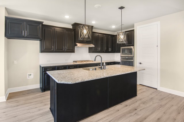 kitchen with stainless steel appliances, light stone countertops, decorative light fixtures, and a center island with sink