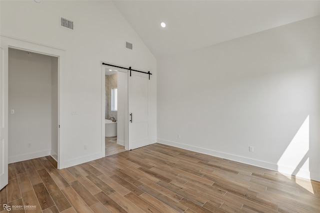 unfurnished bedroom with light hardwood / wood-style flooring, connected bathroom, a barn door, and high vaulted ceiling