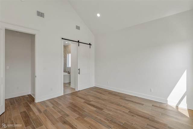 unfurnished bedroom featuring connected bathroom, light hardwood / wood-style flooring, high vaulted ceiling, and a barn door