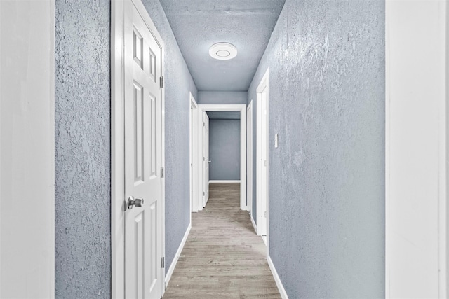 hallway featuring a textured ceiling, a textured wall, light wood-style floors, and baseboards