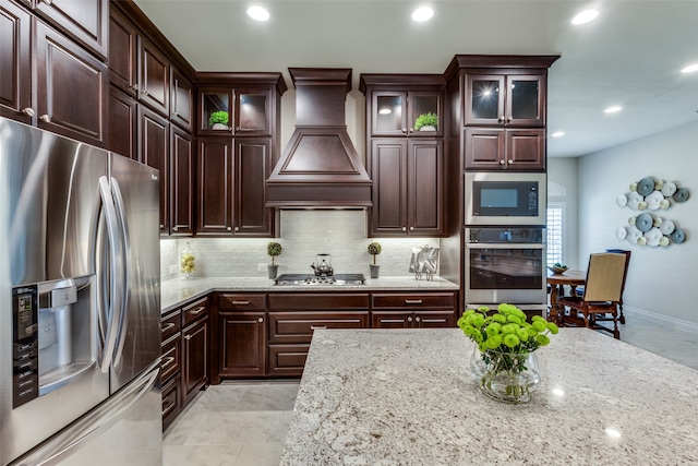 kitchen featuring tasteful backsplash, appliances with stainless steel finishes, light stone countertops, light tile patterned floors, and premium range hood