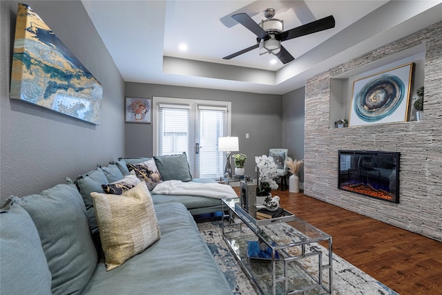 living room featuring ceiling fan, a stone fireplace, a raised ceiling, and dark wood-type flooring