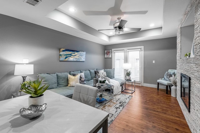 living room with wood-type flooring, ceiling fan, a raised ceiling, and a fireplace