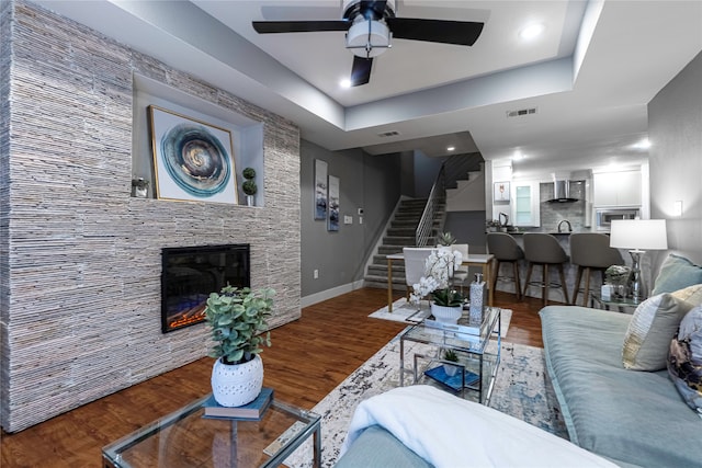 living room with a raised ceiling, a stone fireplace, dark wood-type flooring, and ceiling fan